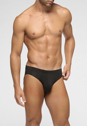 Uomo muscoloso che indossa biancheria intima nera semplice tipo slip, in piedi contro uno sfondo chiaro e semplice.