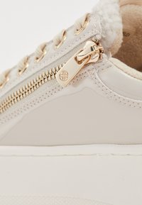 Beige sporta apavi ar spīdīgu lakotu materiālu, zelta aparatūru un kontrastējošu baltu zoli. Iekļauts dekoratīvs rāvējslēdzis un teksturētas detaļas.