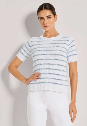 Vrouw met donker haar in een witte kortekorte mouw gebreide trui met blauwe horizontale strepen en een witte broek, poseert met één hand op de heup.