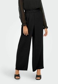 Pantalons larges noirs en tissu texturé avec une taille élastique. Associés à un chemisier noir transparent avec des manches bouffantes.