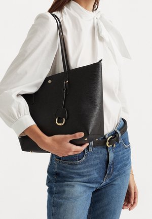 Žena v bílé blůze a modrých džínách drží černou koženou tašku přes rameno se zlatou přezkou a logem.