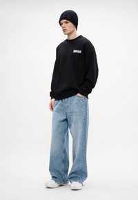 Felpa nera con logo, jeans leggeri blu chiaro a gamba larga e sneakers bianche. Berretto di maglia nera con la parola "HUGO" in lettere in rilievo.