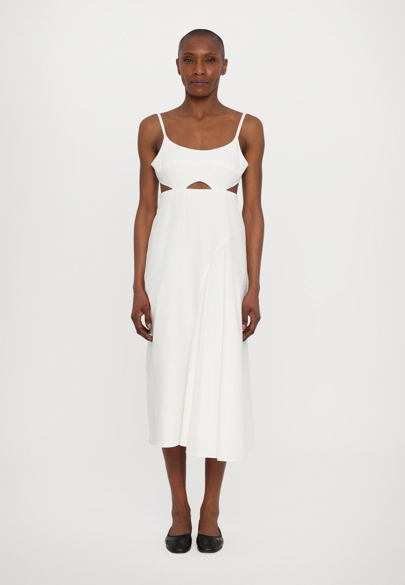 Vestido midi blanco con tirantes finos y un detalle de corte en la cintura. Tejido suave con un ajuste fluido, combinado con zapatos planos negros.