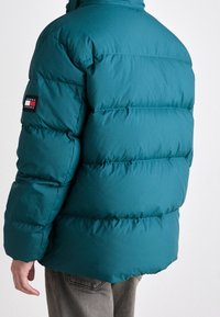 Tyrkysová prošívaná bunda s vodorovným prošíváním, stojáčkovým límcem a nášivkou s logem Tommy Jeans na levém rukávu. Měkká, hladká textilie.