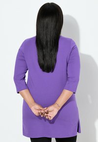 Fioletowa tunika z rękawami do łokcia, bocznymi rozcięciami i luźnym krojem, zestawiona z czarnymi legginsami, pokazana od tyłu z długimi włosami.