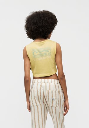 Persoon met krullend haar, gekleed in een geel mouwloos cropped top en lichtgekleurde gestreepte broek, met de rug naar de camera tegen een effen achtergrond.