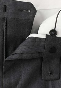 Gros plan de la ceinture d'un pantalon gris foncé montrant une fermeture éclair, un bouton et un crochet avec une doublure en tissu blanc à l’intérieur.