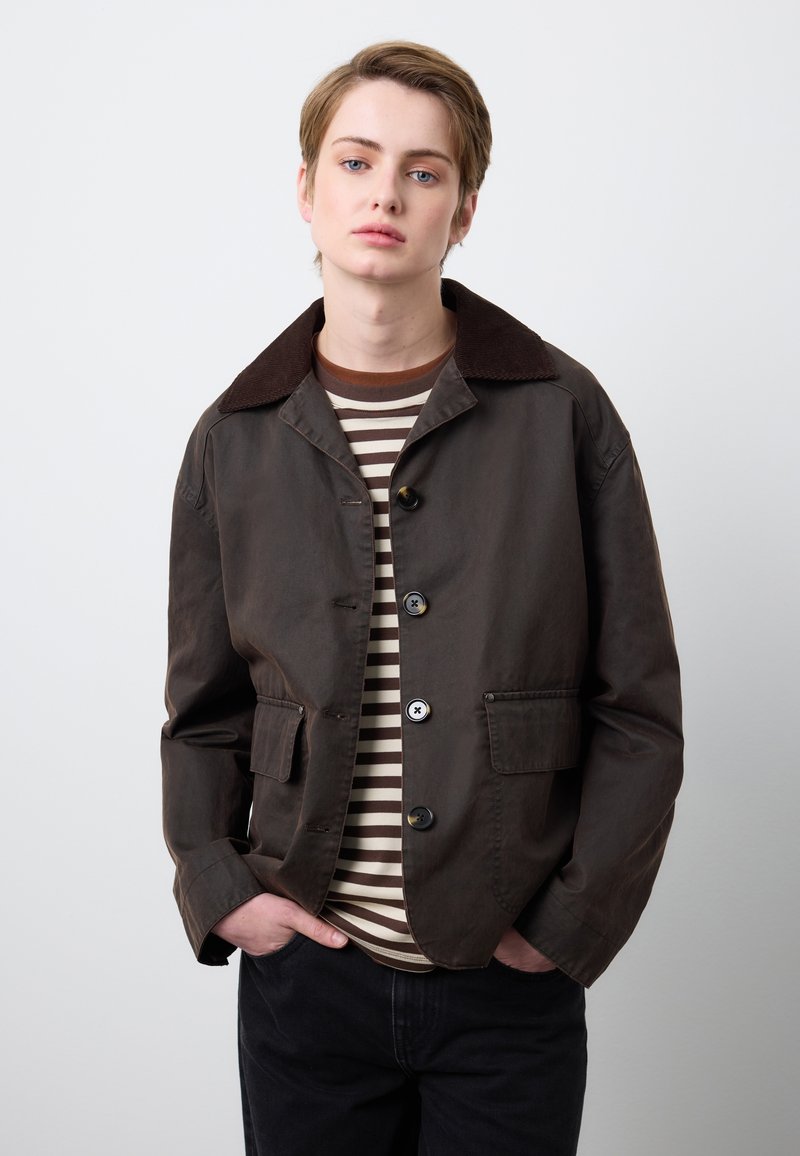 Braune Baumwolljacke mit Cordkragen, ausgestattet mit Knöpfen und zwei Fronttaschen, getragen über einem gestreiften T-Shirt.