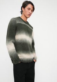 Maglione a righe verde e crema in un morbido tessuto a maglia con polsini e orlo a coste, indossato con pantaloni neri.