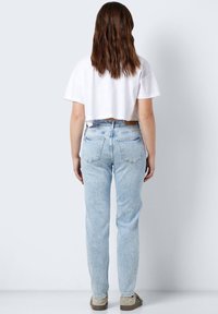 Moteris su ilgais rudais plaukais, vilkinti baltą trumpą marškinėlį, šviesiai mėlynus aukštu liemeniu džinsus ir smėlio spalvos sportbačius, stovinti prieš neutralų foną.