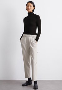 Schwarzer Rollkragenpullover, beiger elastischer Bundhose mit seitlichen Taschen, glatte Textur, getragen mit schwarzen Loafers, vor einem schlichten Hintergrund.