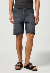 Pantaloni corti in denim scuro con taglio dritto, cinque tasche e dettagli di cucitura in evidenza. Indossati con sandali neri con cinturini regolabili.