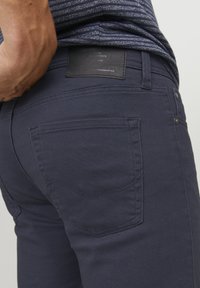 Jeans de mezclilla azul marino con una textura suave, diseño de cinco bolsillos y un parche de cuero negro en la cintura. Detalles de costura sutiles visibles.