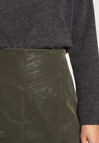 Jupe en cuir vert olive avec une finition texturée, présentant une taille haute structurée et des panneaux en forme de triangle. Pull gris partiellement visible.
