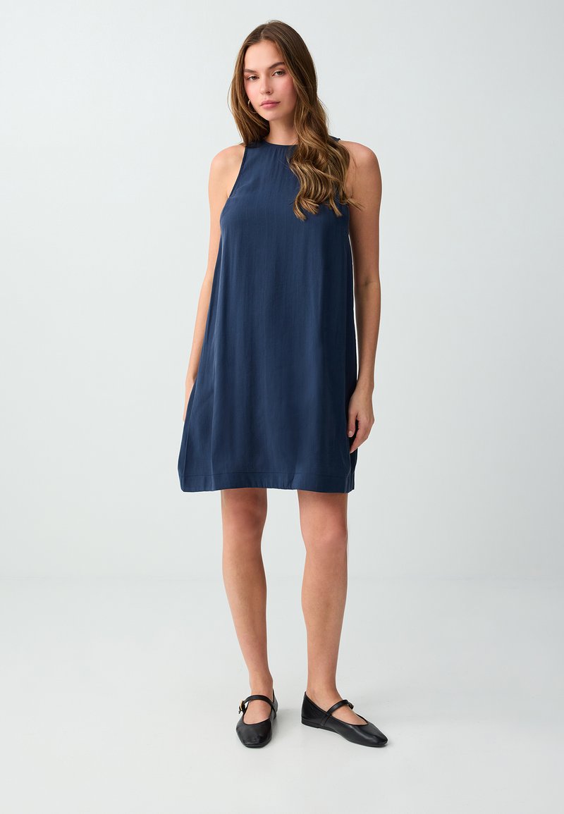 Jeune femme aux longs cheveux bruns portant une robe bleu marine sans manches et des ballerines Mary Jane noires, debout devant un fond clair uni.