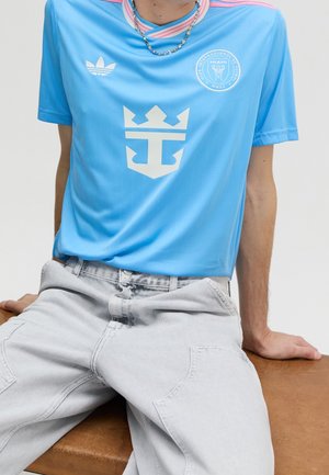Person trägt hellblaues Adidas Fußballtrikot mit Miami Club-Logo und weiße Hose, sitzt auf einer braunen Bank mit den Händen hinter dem Rücken.