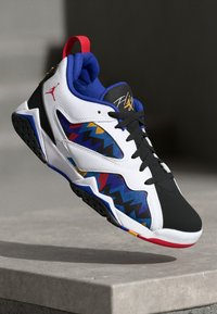 Sneaker Air Jordan bianco, nero e blu con motivi zigzag multicolori, con un'etichetta rossa e lacci neri, sospesa sopra una superficie di cemento.