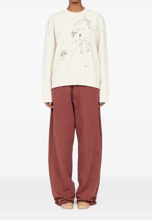 Persona che indossa pantaloni marroni larghi, scarpe beige e una felpa color crema con un disegno a linee di un cavallo rampante con un grande fiocco sopra.