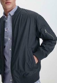 Marineblaue Bomberjacke mit einer Reißverschluss-Tasche am linken Arm, gerippten Ärmelbündchen und einem glatten, leichten Stoff. Gestreiftes Hemd darunter.