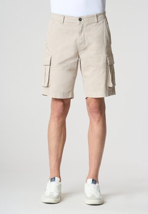 Homme portant un short cargo beige avec des poches latérales et des baskets blanches, debout devant un fond clair uni.