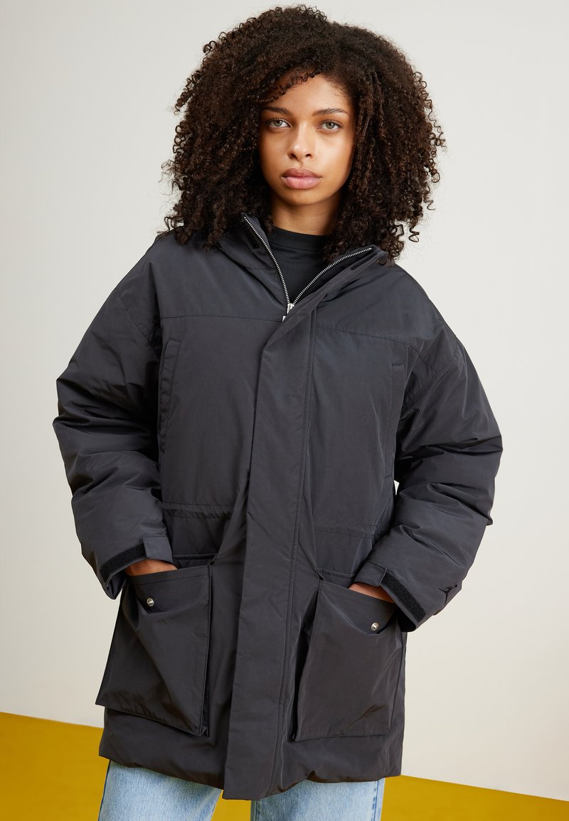 Veste rembourrée noire avec un col haut, fermeture éclair et grandes poches avant. Texture lisse avec une coupe décontractée et des manches à revers.
