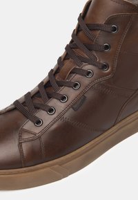 Scarpa alta in pelle marrone con superficie testurizzata, punta rotonda e lacci marroni. Presenta occhielli scuri e una suola in gomma color cuoio.