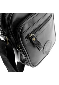 Bolso de hombro negro de cuero con superficie texturizada, compartimentos con cremallera y una etiqueta decorativa con un acento de latón. Incluye correa ajustable.