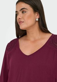 Bordeauxrote Bluse mit V-Ausschnitt und filigranen Spitzenakzenten auf den Schultern. Hergestellt aus weichem, strukturiertem Stoff.