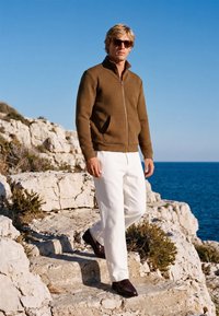 Homme blond portant une veste marron zippée, un pantalon blanc et des chaussures foncées marchant sur des falaises rocheuses au bord d’une mer bleue sous un ciel dégagé.