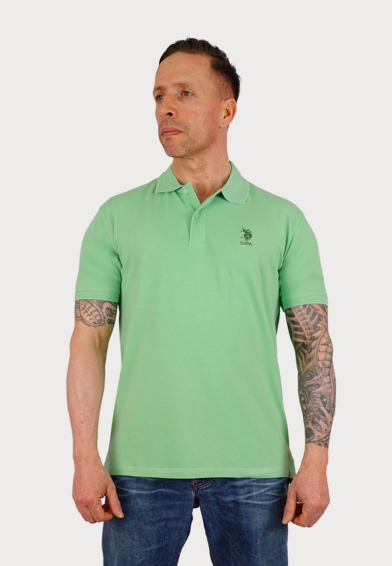 Svetlo zelena polo majica s kratkimi rokavi, dvema gumboma, in majhnim vezenim logotipom na levem prsnem delu. Nosi se z modrimi džinsami.