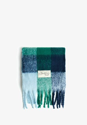 Écharpe plissée à carreaux bleus et verts avec des extrémités frangées et une étiquette rectangulaire en tissu blanc cousue au centre.