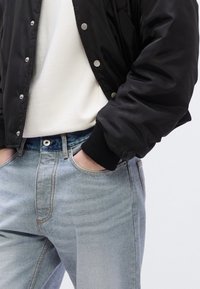 Μαύρο μπουφάν τύπου μπομπέρ με κουμπιά πατιλέτας, λευκό μπλουζάκι και ανοιχτόχρωμα τζιν με ξεθωριασμένο πλύσιμο. Ορατές τσέπες και ριμπ μανσέτες στο μπουφάν.