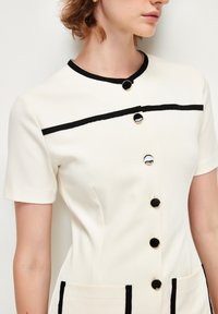 Das weiße Kurzarmkleid hat schwarze Akzente entlang des Kragens und der Taschen, große runde schwarze Knöpfe und ein strukturiertes Design.