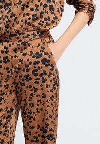 La mano de una mujer en el bolsillo de unos pantalones de satén marrones con estampado de leopardo negro, con la manga de la camisa a juego enrollada, contra un fondo claro.
