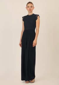 Mono de encaje azul marino, con mangas cortas y un torso ajustado, pantalones de pierna ancha fluidos y sandalias plateadas elegantes.