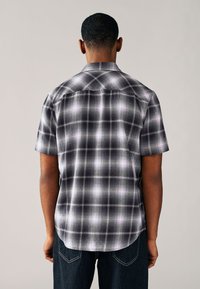 Kortärmad rutmönstrad skjorta i svart och grått, med knappslå vid kragen, slät textur och rundad fåll. Bakifrån syns subtila mönsterdetaljer.