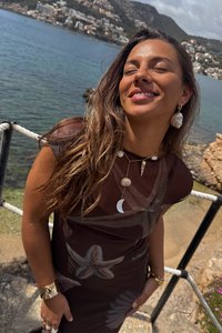 Vestido castanho transparente com padrões de estrela-do-mar, sobreposto com várias colares que apresentam conchas e contas. Cabelo longo e ondulado, acessórios mínimos.
