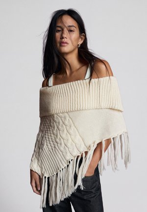 Femme aux longs cheveux noirs porte un poncho en maille crème, épaules dénudées, avec des franges, et un pantalon noir sur un fond gris uni.