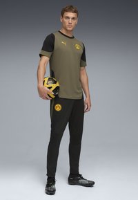 Puma sportstøjssæt med en olivengrøn skjorte med sorte ærmer, sorte bukser og en sort og gul fodbold.