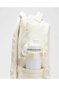 Mochila beige con una superficie texturizada, que cuenta con un bolsillo lateral que sostiene una botella de agua blanca con una tapa de rosca gris y correa.