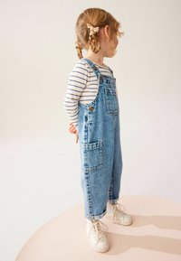 Mono de mezclilla con tirantes ajustables, puños enrollados y bolsillos frontales. Lleva puesto una camiseta de rayas de manga larga y está combinado con zapatillas blancas.