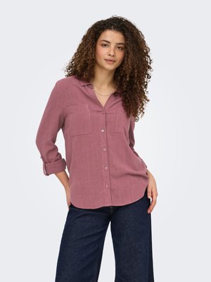 Junge Frau mit lockigem Haar, die ein altviolettes Hemd mit hochgekrempelten Ärmeln und dunkle, weite Jeans trägt und vor einem hellen Hintergrund steht.