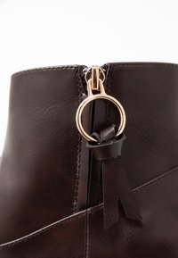 Botín de cuero marrón que presenta un zipper dorado, detalle de anilla para tirar y una superficie texturizada con una costura en un lado.