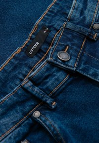 Vaqueros de mezclilla en azul oscuro con costuras en marrón que contrastan. Presenta una etiqueta negra y herrajes de botón plateado cerca de la cintura.