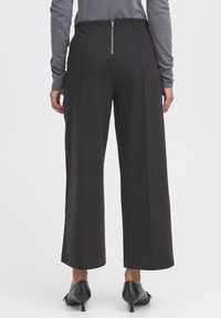 Tył osoby noszącej czarne spodnie z wysokim stanem i szerokimi nogawkami z widocznym tylnym zamkiem, szary długi rękaw oraz czarne buty na obcasie.