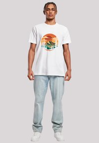 White cotton t-shirt featuring a circular graphic with mountains, trees, and a sunset in orange and green tones. Paired with light denim jeans.