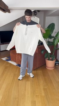 Weißes oversized Sweatshirt mit Reißverschlusskragen, gerippten Bündchen und Seitentaschen. Das Modell hält es vor einem lichtdurchfluteten Raum mit Pflanzen hoch.