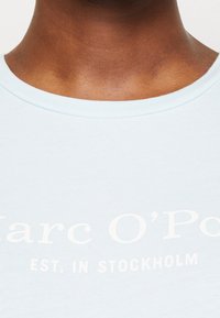Camiseta de algodón azul claro con cuello redondo y texto blanco que dice "Marc O'Polo EST. EN ESTOCOLMO." Textura suave, diseño ajustado.