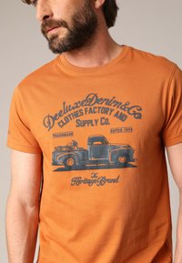 Oranje katoenen T-shirt met een blauwe afbeelding van een vintage vrachtwagen, met de tekst "Deeluxe Denim & Co." en daaronder "The Heritage Brand".