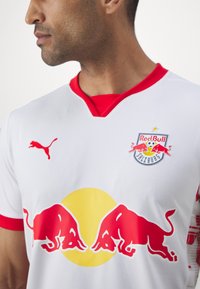 Bijela majica s crvenim akcentima, s dva crvena bika i žutom kružnicom. Puma logo i grb Red Bull Salzburga na prsima.